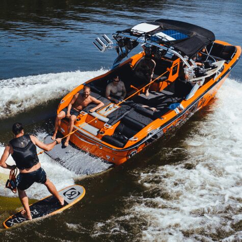 Person wakesurfs behind a sleek black and orange speedboat on a sunny day.